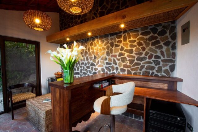 Cozy workspace featuring a wooden desk, potted white flowers, a wicker chair, and stone wall decor, illuminated by stylish pendant lights.