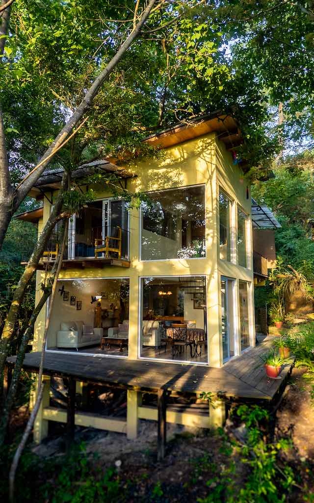 Two-story modern house with large glass windows and a deck, surrounded by dense green trees and sunlight filtering through the leaves.