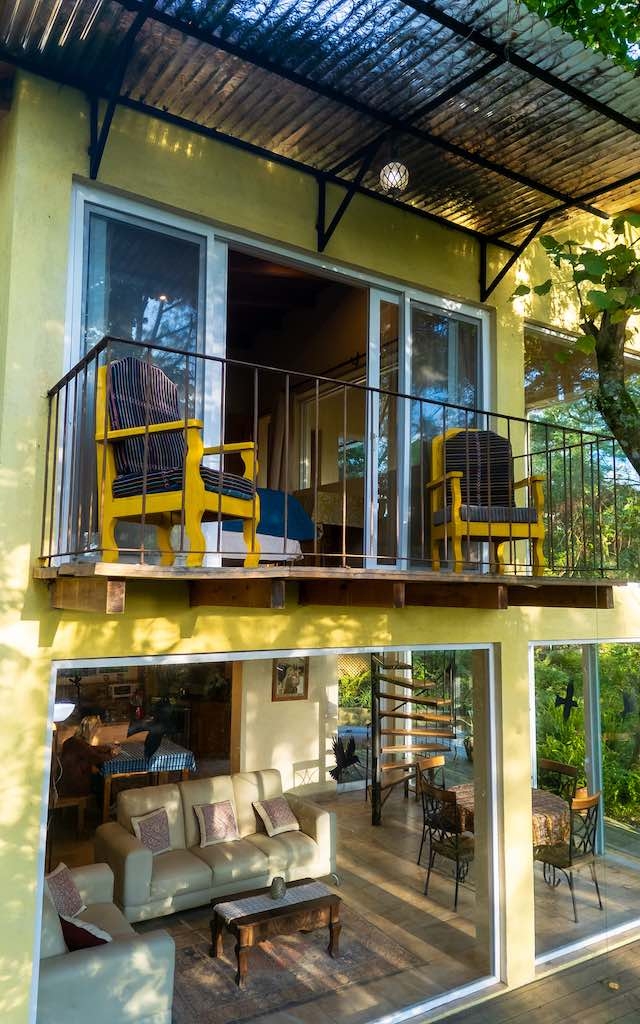 A vibrant yellow house with large windows, featuring a balcony with yellow chairs and a cozy living area visible through the glass.