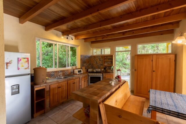 A cozy kitchen featuring wooden cabinets, a dining table, and large windows overlooking greenery, with modern appliances and natural light.