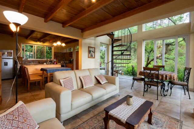 Cozy living space featuring a light sofa, dining area, spiral staircase, and large windows with a lush outdoor view.