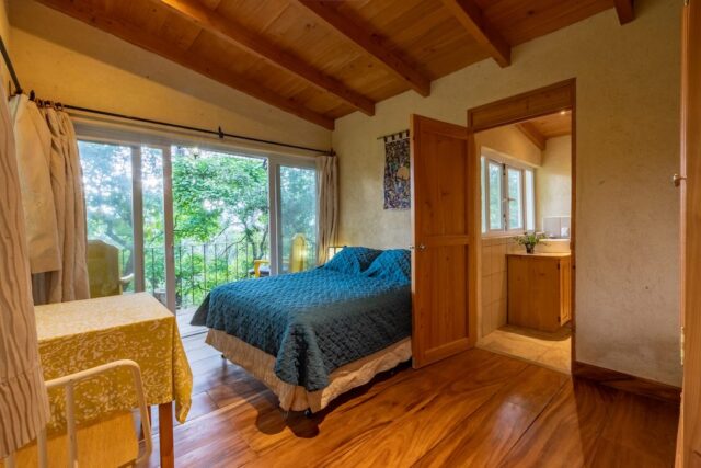 Cozy bedroom with a wooden ceiling, a blue quilted bed, and large windows overlooking greenery, connected to a small bathroom.